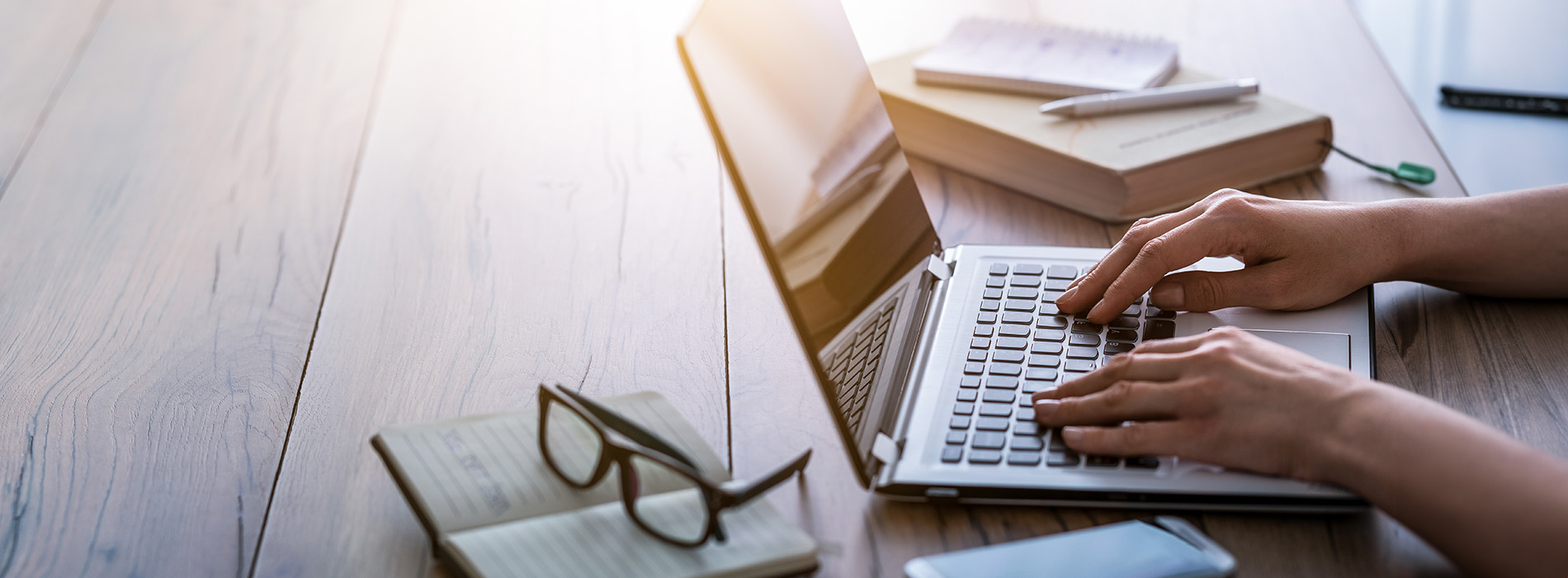 A person typing on a laptop at a desk with books and papers, set against a blurred background of a coffee table and a window with natural light.