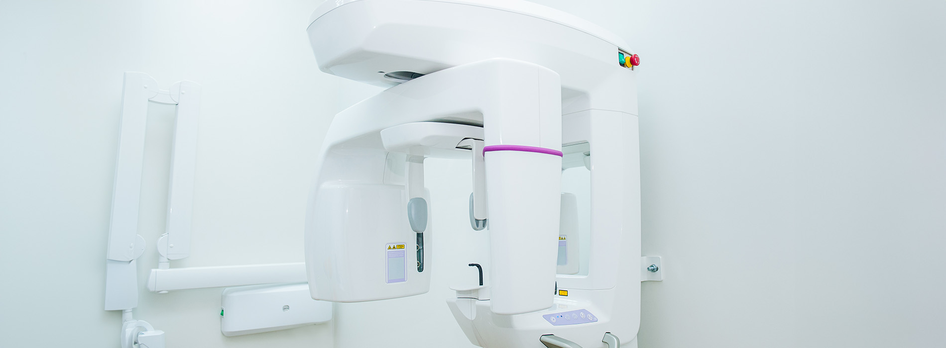 The image shows a modern medical imaging machine, specifically an MRI scanner, with a purple interior visible through its transparent door, situated inside a room with white walls and a white ceiling.