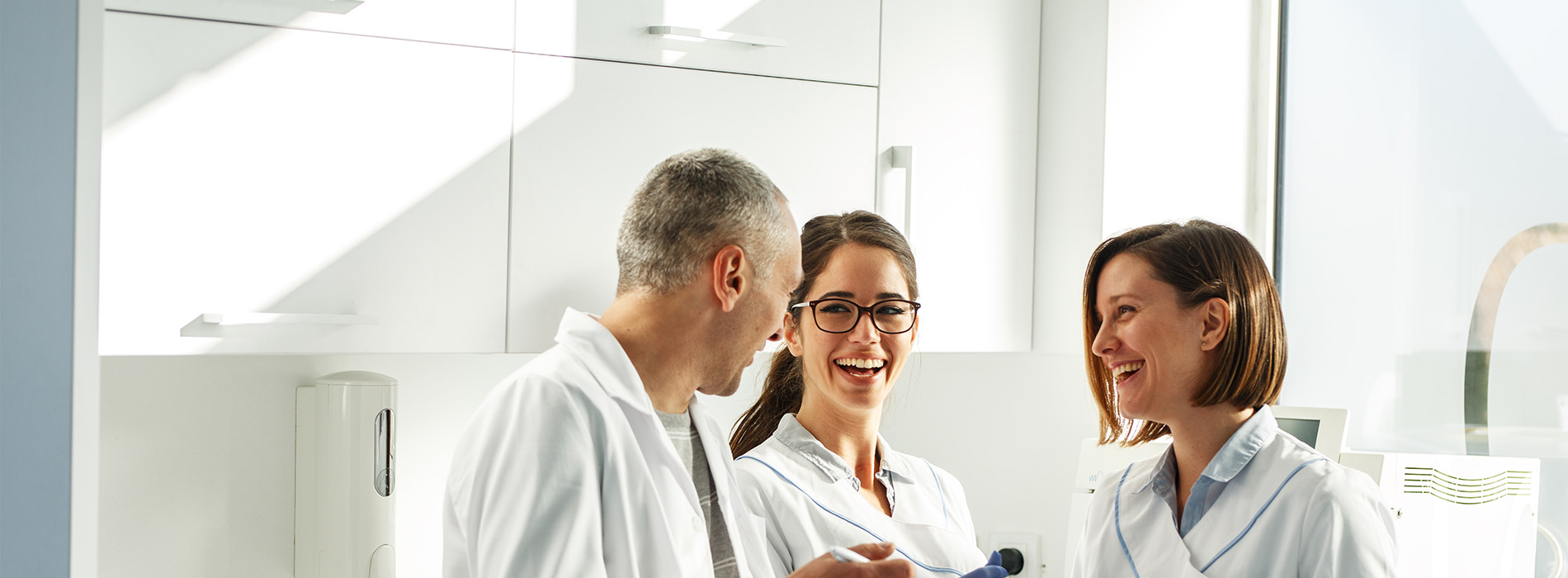 The image shows three individuals in a professional setting, likely an office or laboratory, with two standing and one seated behind them. They appear to be engaged in conversation, possibly discussing work-related matters.