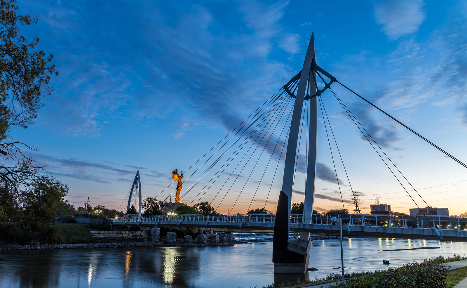 The image depicts a serene nighttime scene at a bridge with a view of a river and a city skyline, featuring a suspension bridge with a lit-up tower in the background and a tranquil waterfront setting.