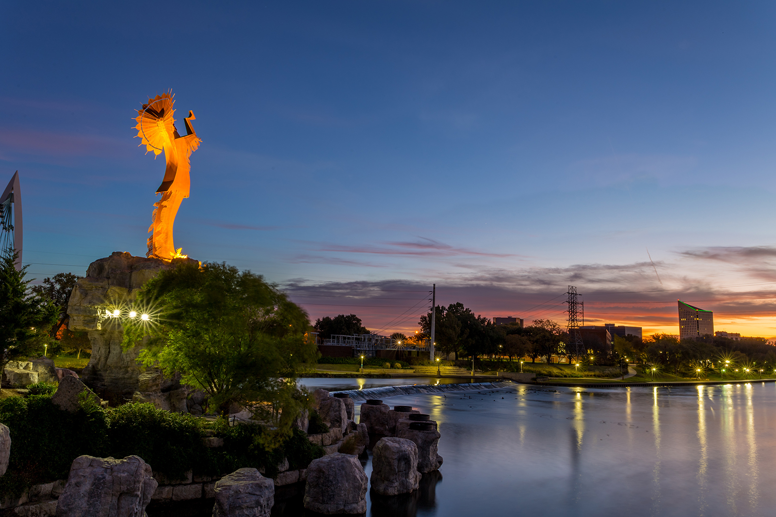 The image depicts a statue at sunset with a body of water in the foreground, under a clear sky.
