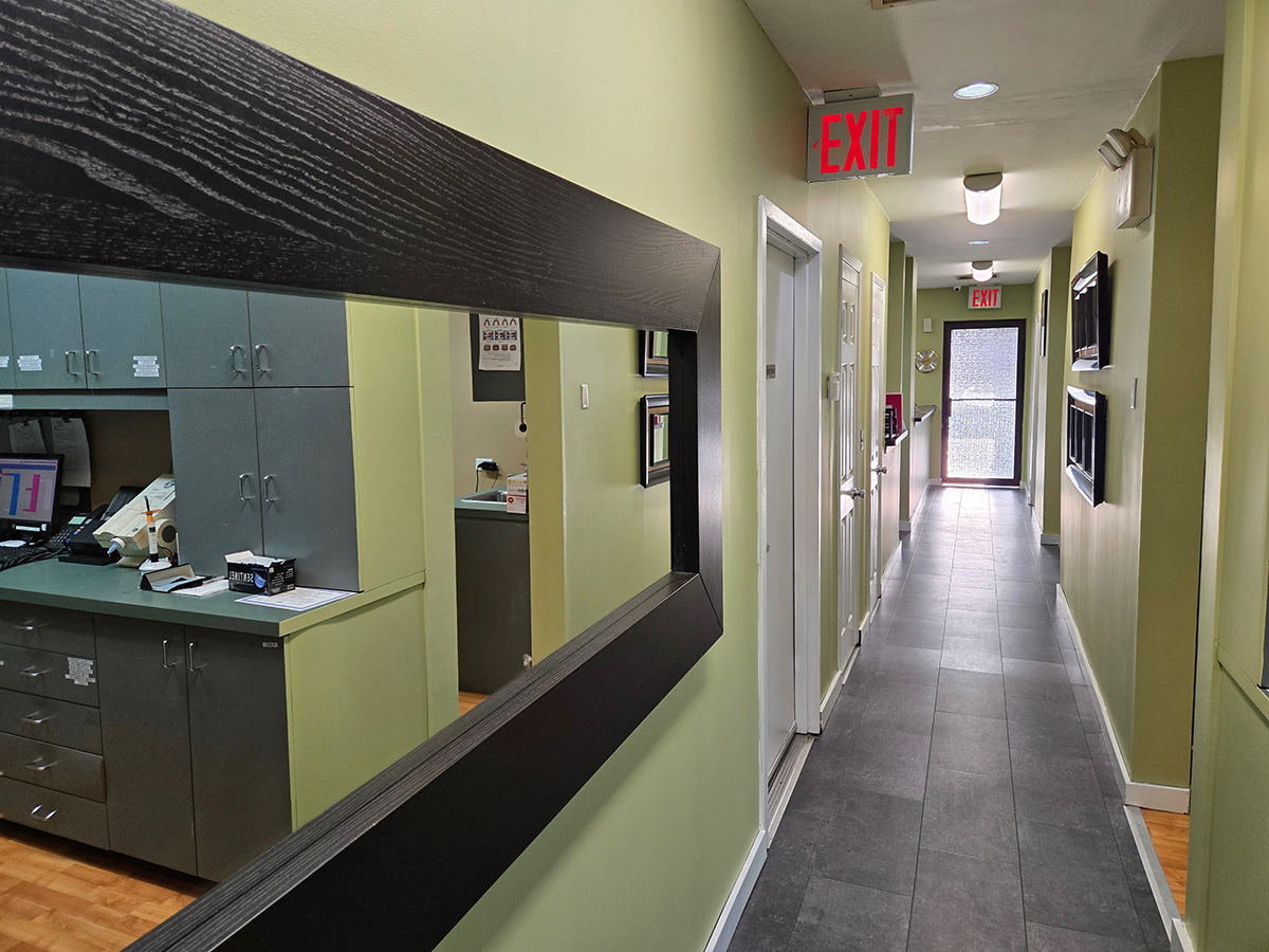 The image depicts an interior view of a well-lit office space with a modern design, featuring a reception area, a hallway leading to other rooms, and a waiting area with a desk and chairs visible through a mirror reflection.