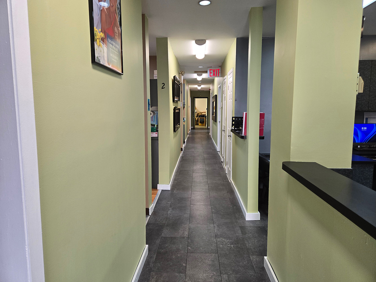 The image shows an interior hallway with a long corridor leading to a doorway on the right side. The walls are painted yellow, and there's a green accent wall at the end of the hall. The flooring appears to be tile, and there's a countertop with a black surface on the left side of the photo.