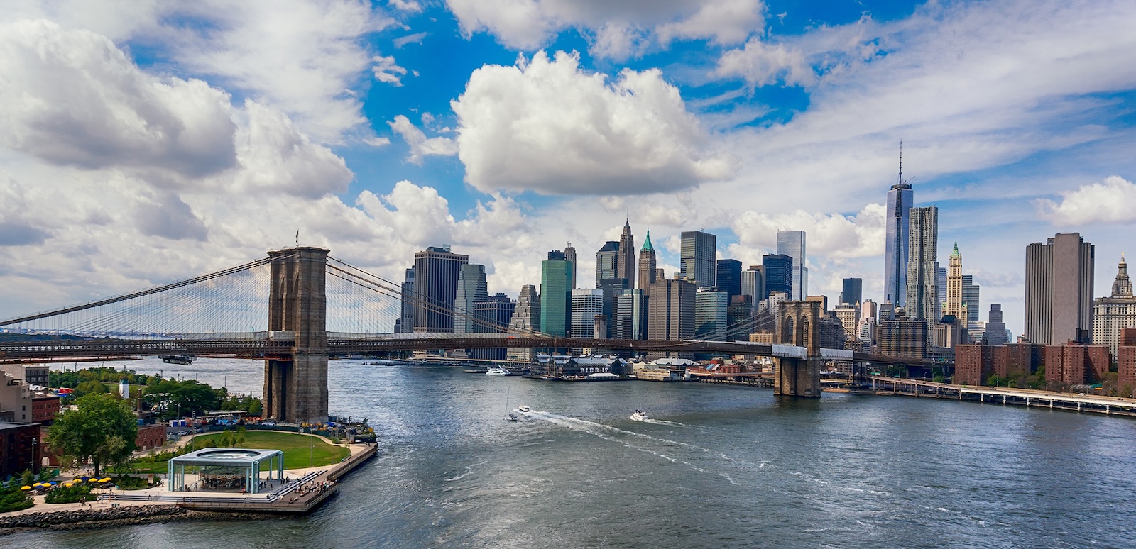 The image depicts a city skyline with iconic landmarks such as the Brooklyn Bridge and the Statue of Liberty, viewed from a vantage point across a river, under a partly cloudy sky.