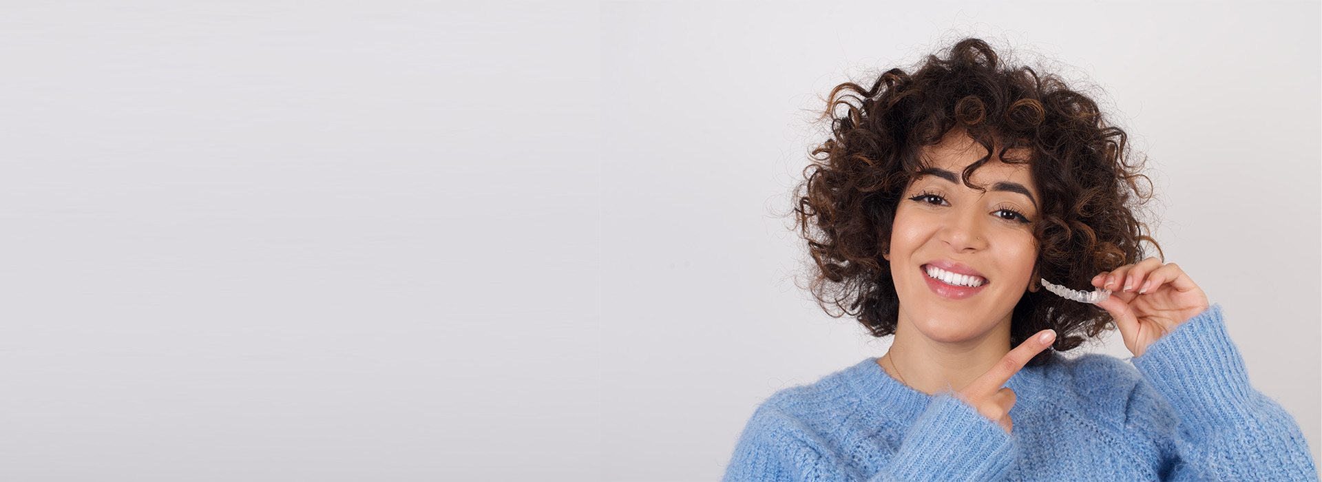 A woman with short curly hair smiles at the camera while holding up her phone, set against a plain background.