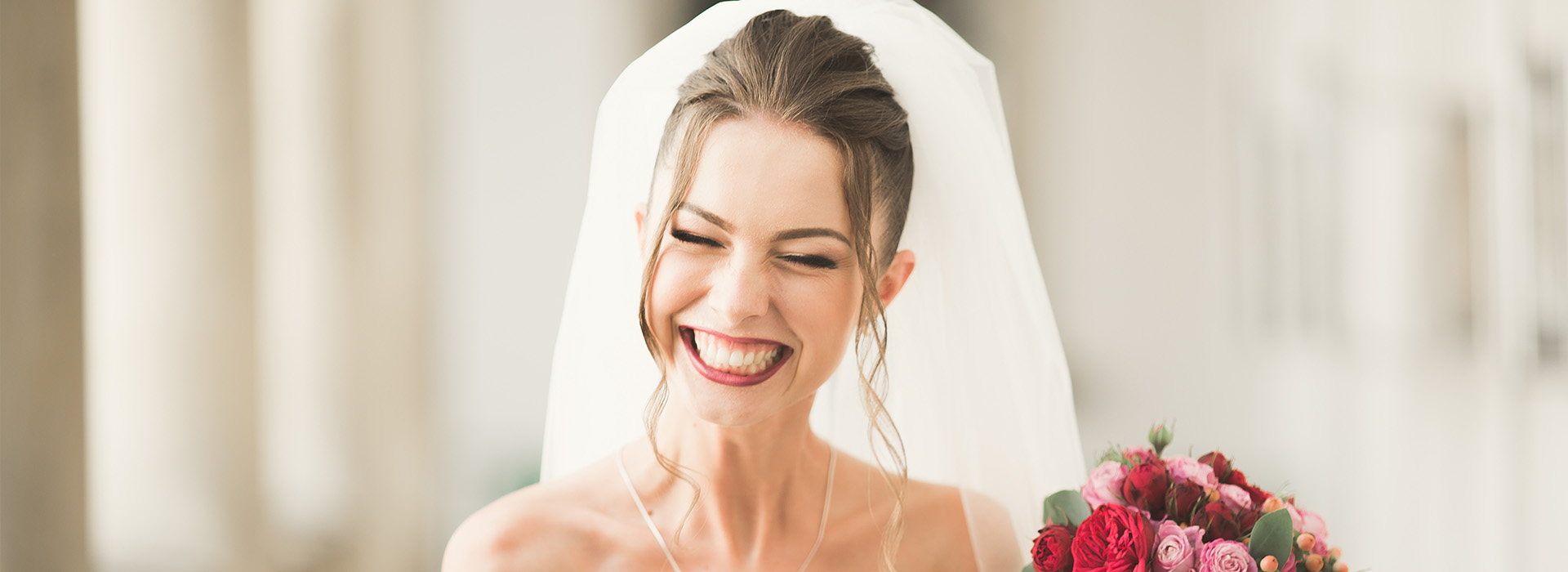 A bride wearing a traditional white wedding dress with her mouth open, laughing, and holding a bouquet, smiling at the camera.