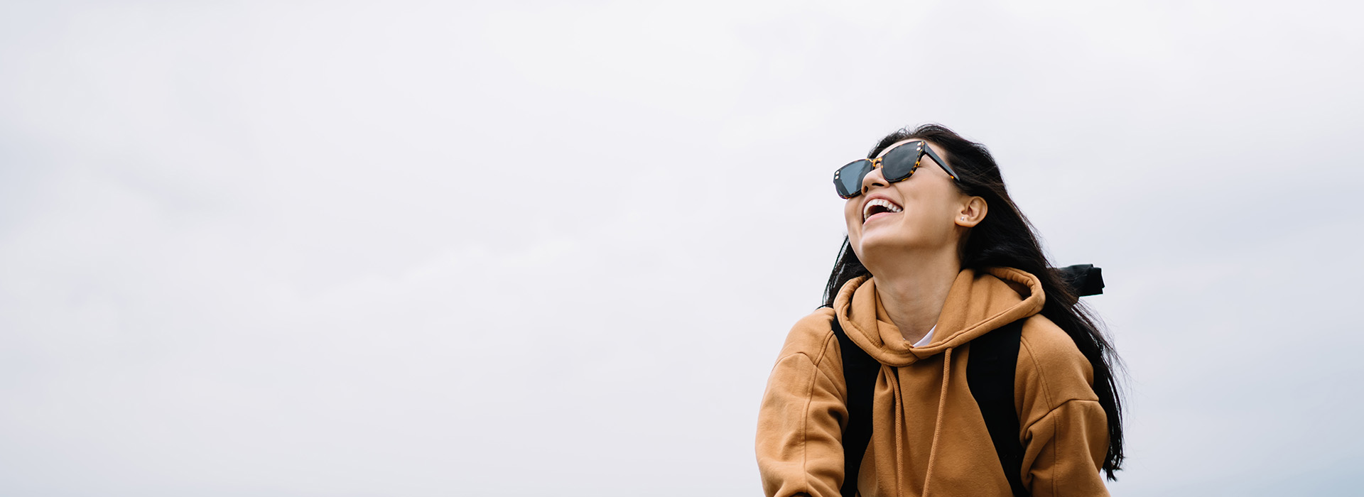 The image shows a person standing outdoors with sunglasses on, wearing a backpack and a hoodie, smiling at the camera, against a cloudy sky background.