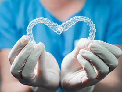 The image shows a hand holding a clear dental aligner shaped like a heart, with a person wearing medical gloves in the background.