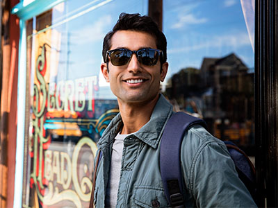 A man wearing sunglasses and a backpack stands outside a storefront, posing with a smile for the camera.