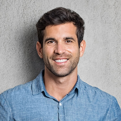 A man with a beard and short hair is smiling at the camera, wearing a light blue shirt and standing against a concrete wall.