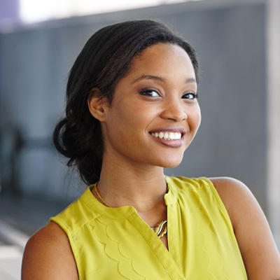 A young woman with a radiant smile, posing confidently against a blurred background, wearing a yellow top and smiling at the camera.