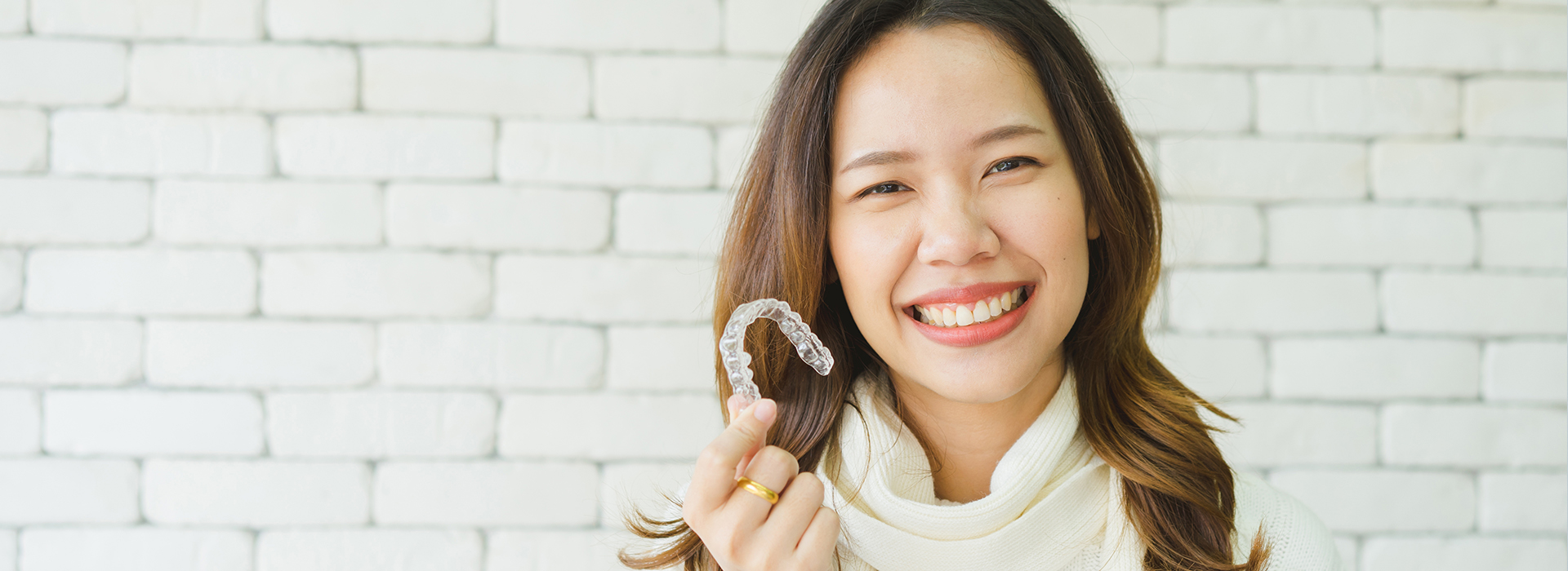 A woman with a smile, wearing a necklace and holding a ring, against a brick wall background.