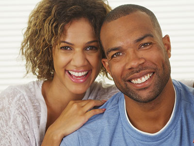 The image shows a smiling man and woman posing together with the man having his arm around the woman s shoulder, both appear happy and are looking directly at the camera.