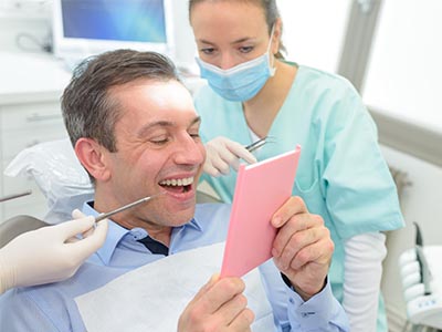 A man sitting in a dentist s chair, smiling at a pink card he holds, with a woman dental hygienist standing behind him examining his teeth.