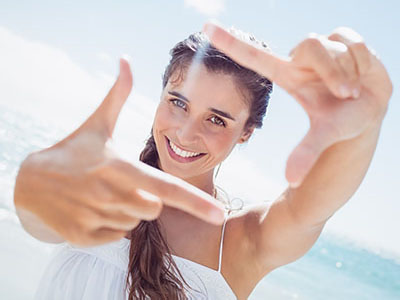 The image shows a woman with long hair smiling at the camera while holding her finger up against the lens, creating a framing effect with her hand.