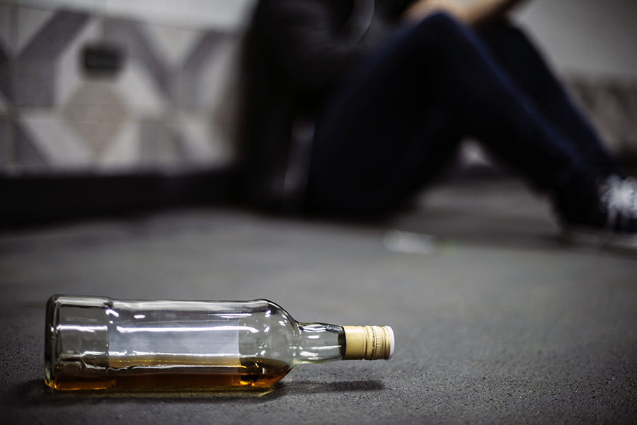 alcoholism concept - whiskey bottle laying nearby a person sitting on the floor out of focus - Oral dysplasia and cancer on the rise with COVID-related alcohol consumption